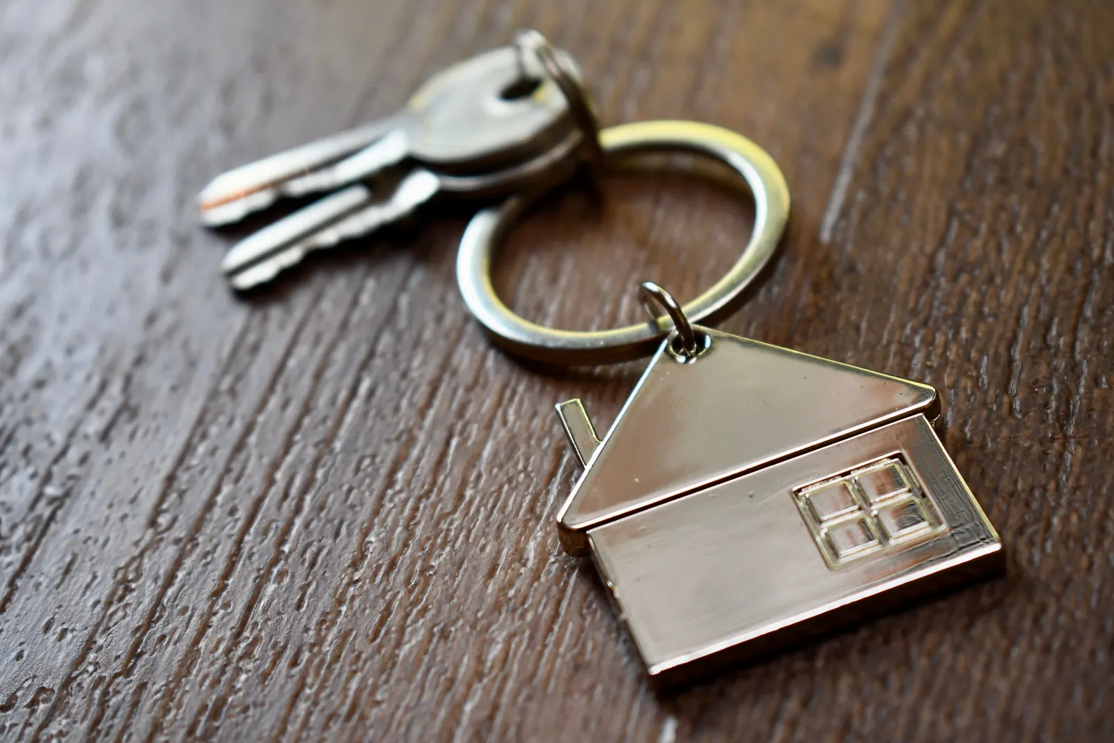 Two keys on a ring with a silver house-shaped key chain, lying on a dark wooden table