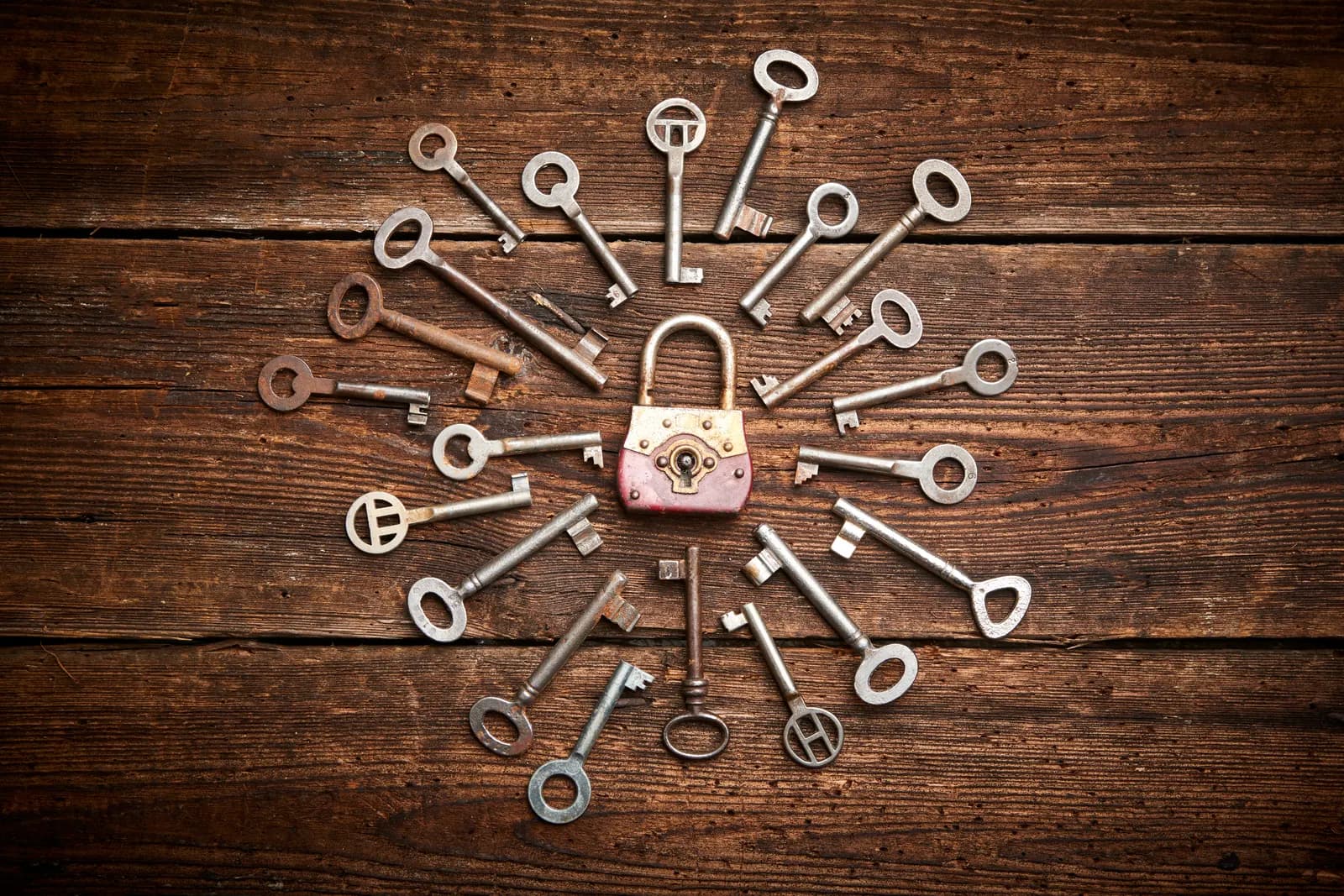 An old padlock on a wooden table, surrounded by a ring of antique keys pointing toward it