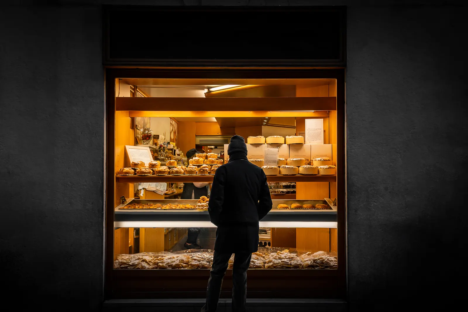 A person in a dark coat and knit cap stands at night in front of a lit bakery window full of pastries
