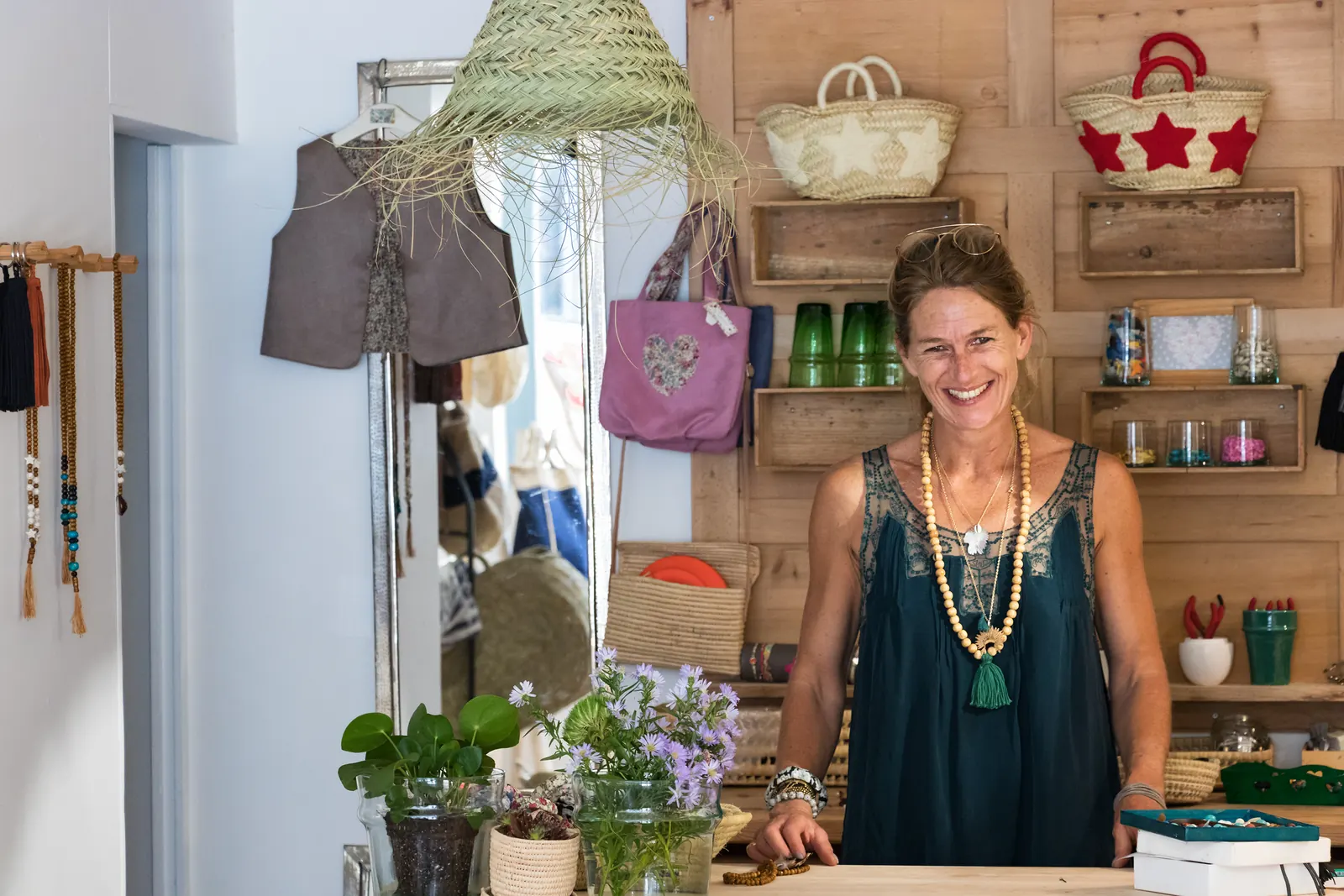 A smiling shop owner in a long wooden-bead necklace stands behind the counter of her small gift shop, with baskets and bags on wooden shelves behind her