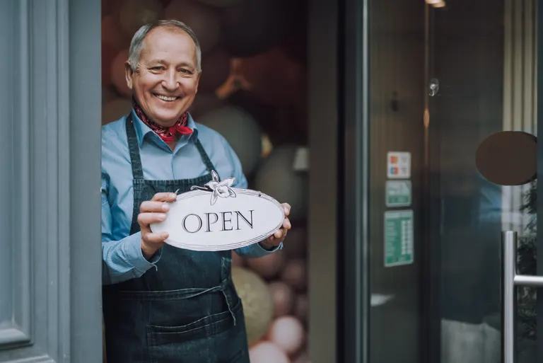 A smiling shop owner in an apron holds an Open sign in the doorway of his cafe