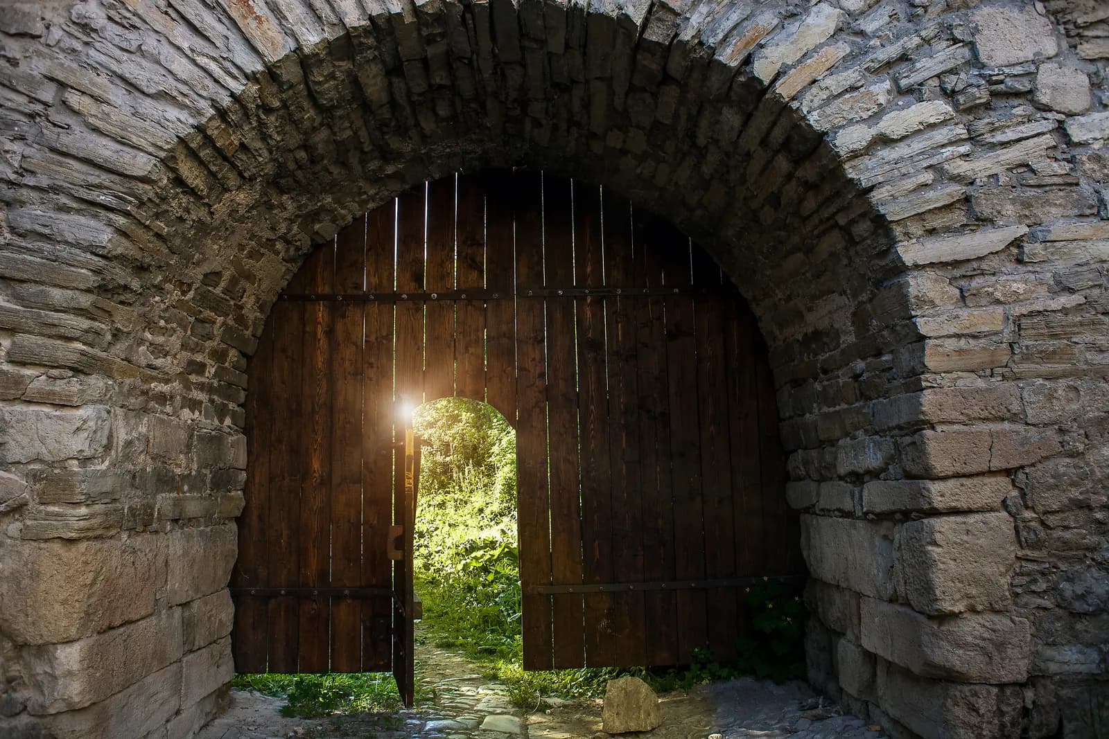 A tall wooden gate in an old stone archway, with a small door in it standing open onto a sunlit garden path