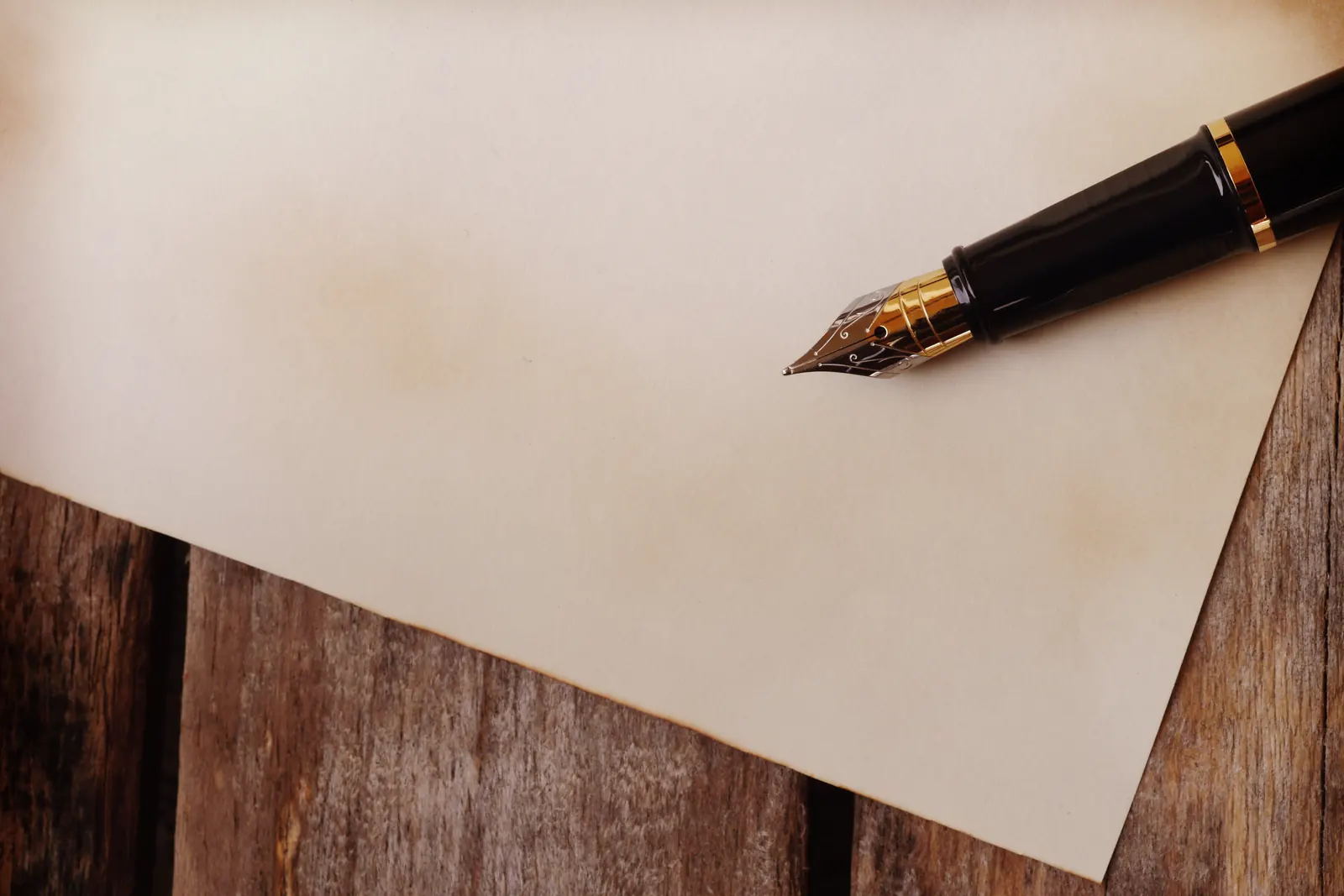 A black fountain pen with a gold nib resting on a blank sheet of aged paper on a wooden table