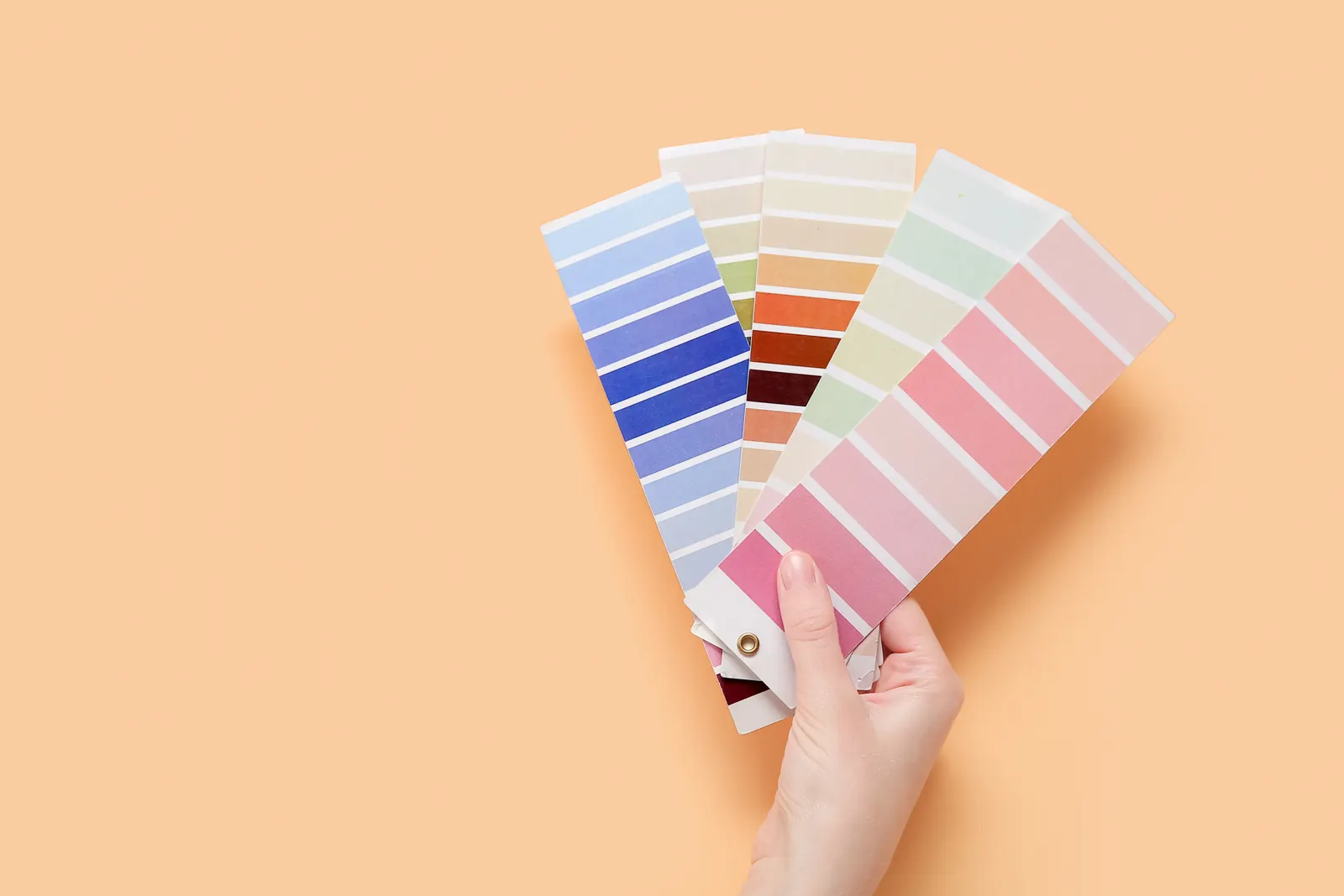 A hand holding a fan of paint color swatches against a peach background