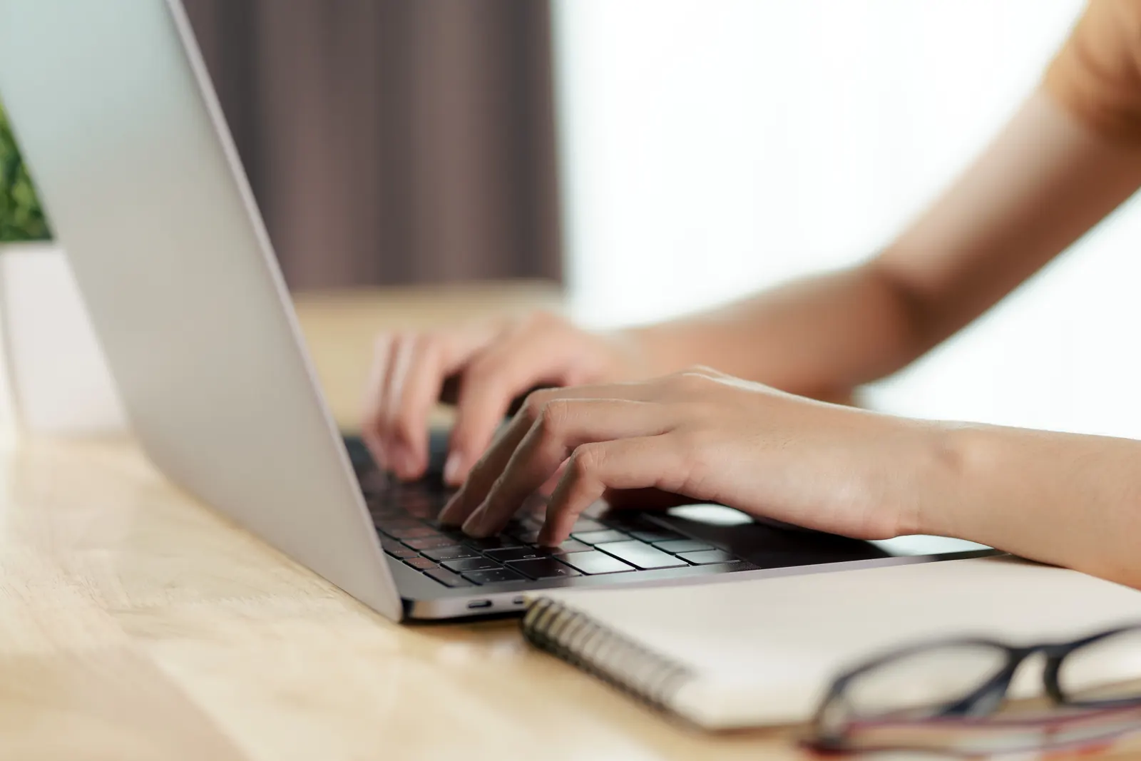 Hands typing on a laptop on a wooden desk, beside a notebook and a pair of glasses