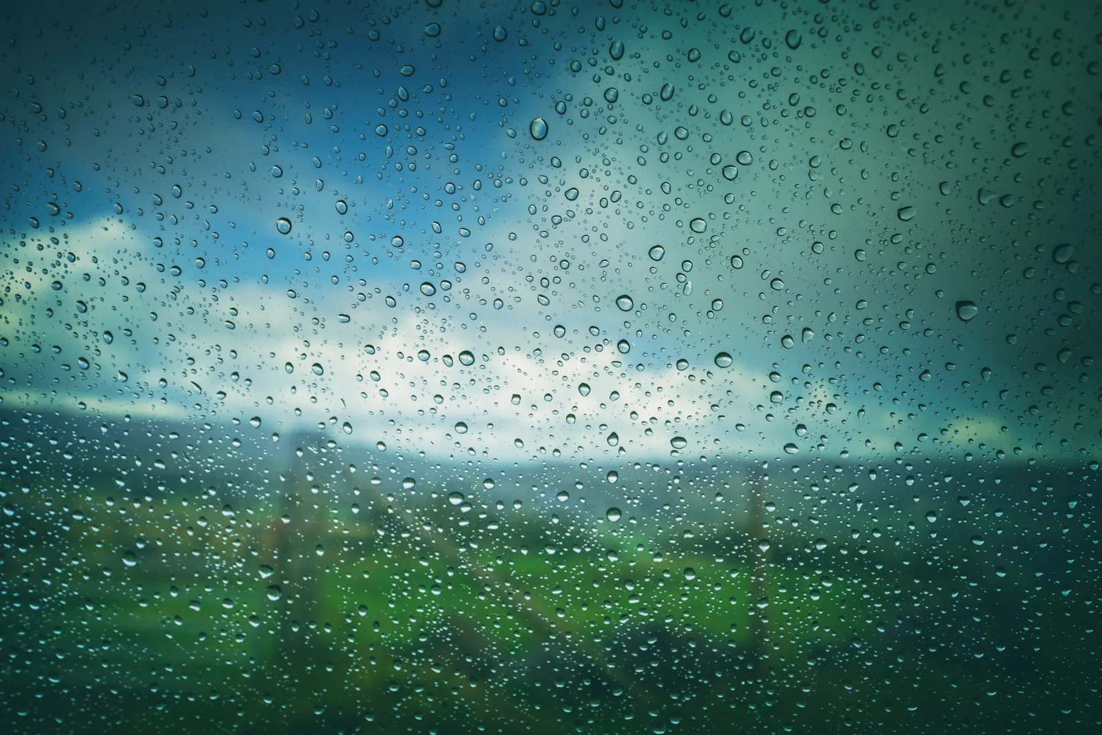 Raindrops on a car window, with green fields and a cloudy sky blurred behind the glass
