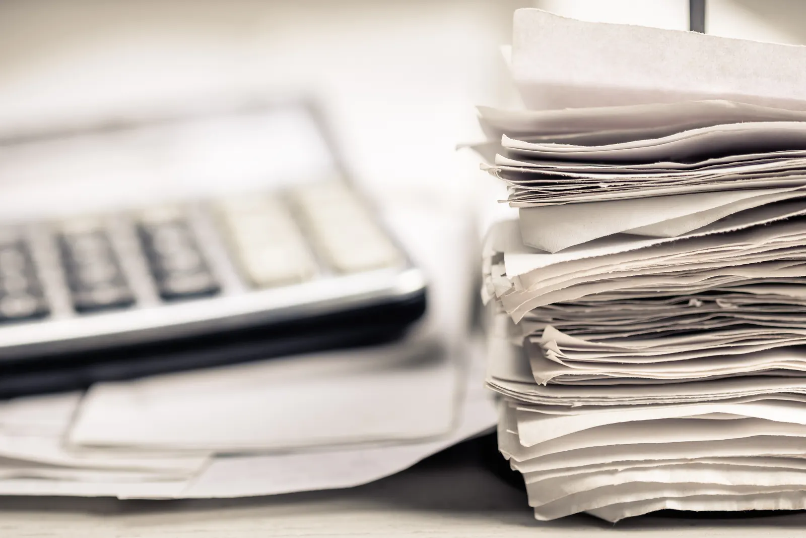 A tall stack of paper receipts on a spike beside an out-of-focus calculator on a desk