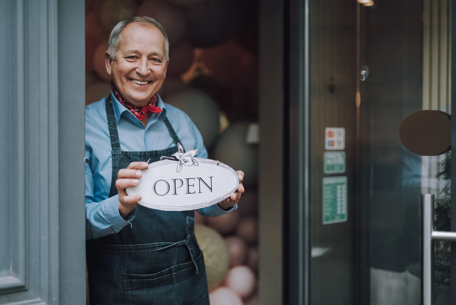 A smiling shop owner in an apron holds an Open sign in the doorway of his cafe
