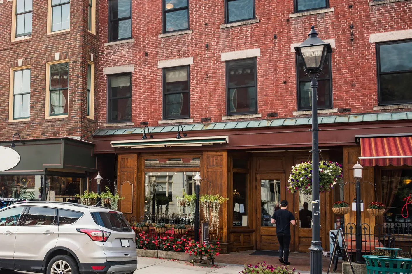 A wood-fronted shop in a brick building on a small-town main street, with hanging flower baskets and a customer walking to the door