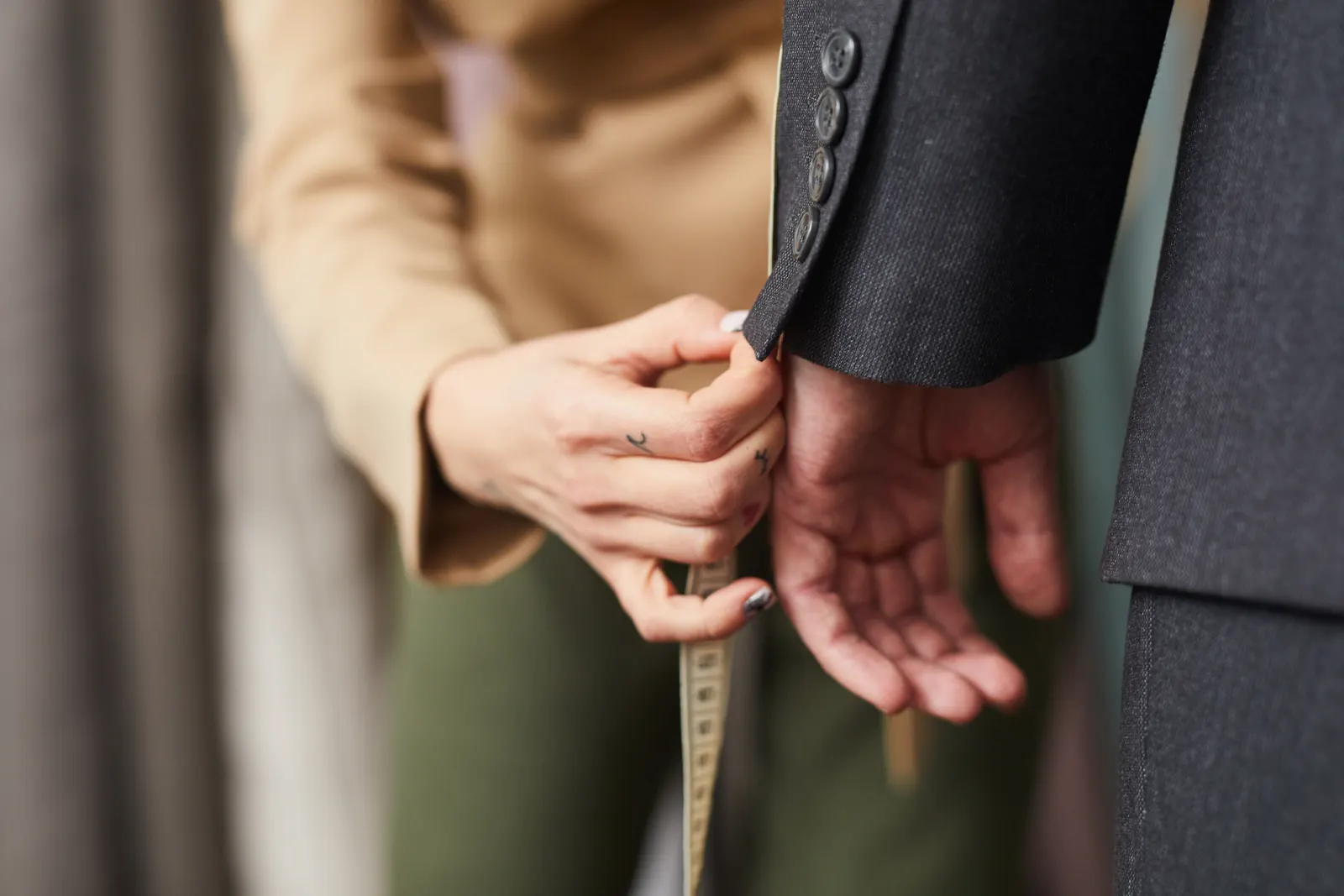 A tailor's hands hold a tape measure at the cuff of a client's dark suit sleeve